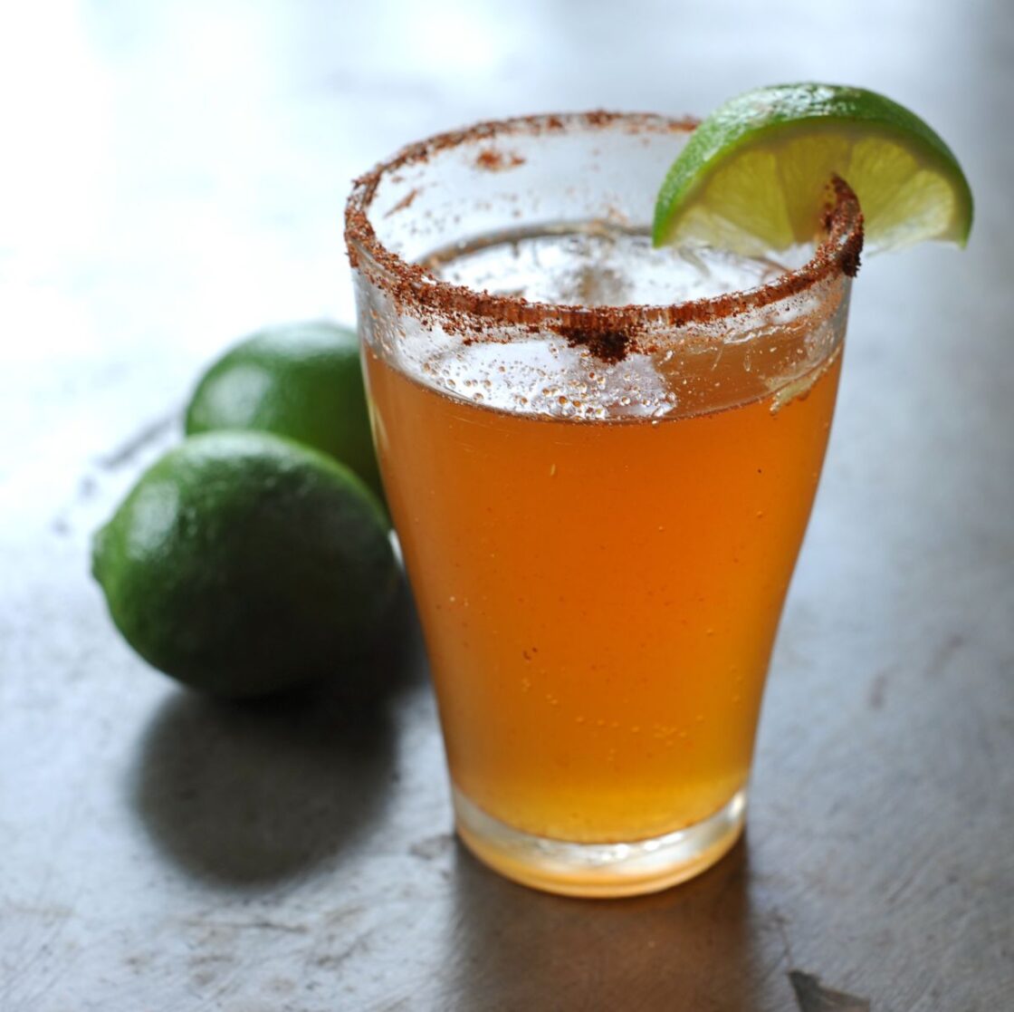 Image: clear glass with michelada beer cocktail with chili salted rim and sliced lime garnish, two limes in background on slate table. a michelada recipe is easy to make and super refreshing via Organic Authority.