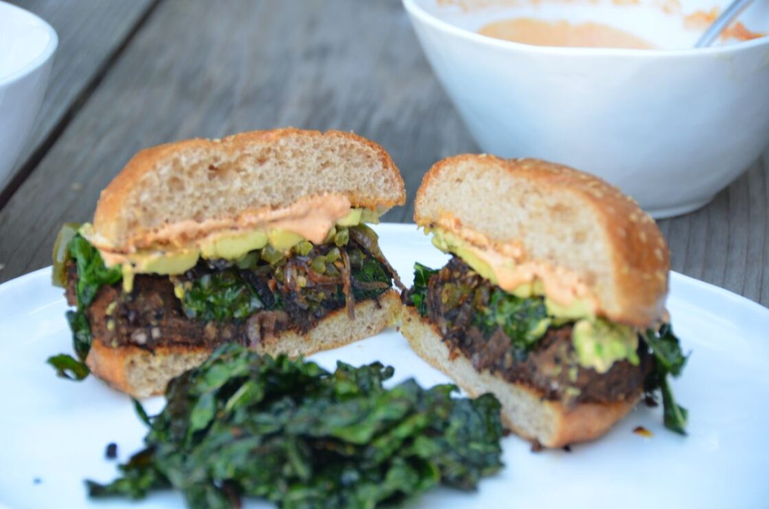 Image: Black Bean veggie Burgers with Caramelized Onions, Jalapeños, Avocado and Blistered Kale on a white plate and white bowl with sauce in the background. These can be made as vegan black bean burgers via Organic Authority