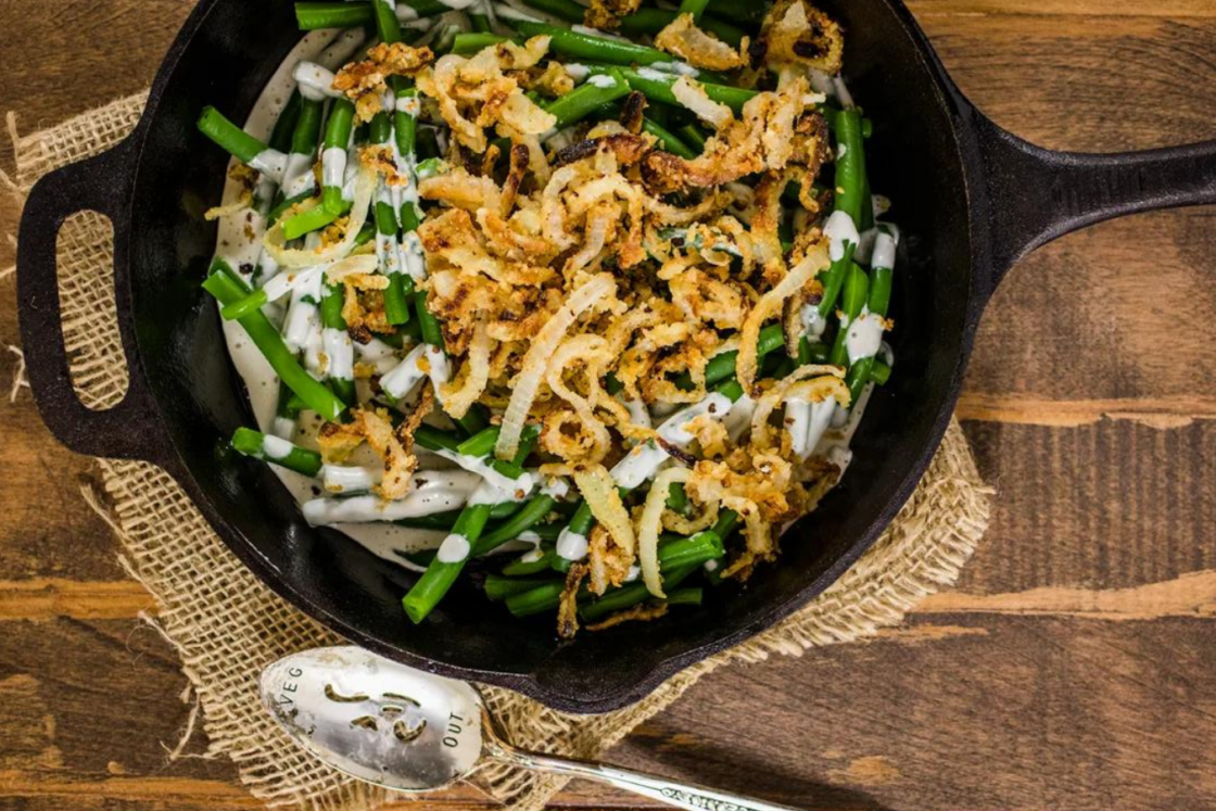 Image of homemade vegan green bean casserole in an iron skillet on top of a small piece of burlap, with a silver slotted spoon laying beside on a wood work surface showing how to make fresh green beans into a casserole.