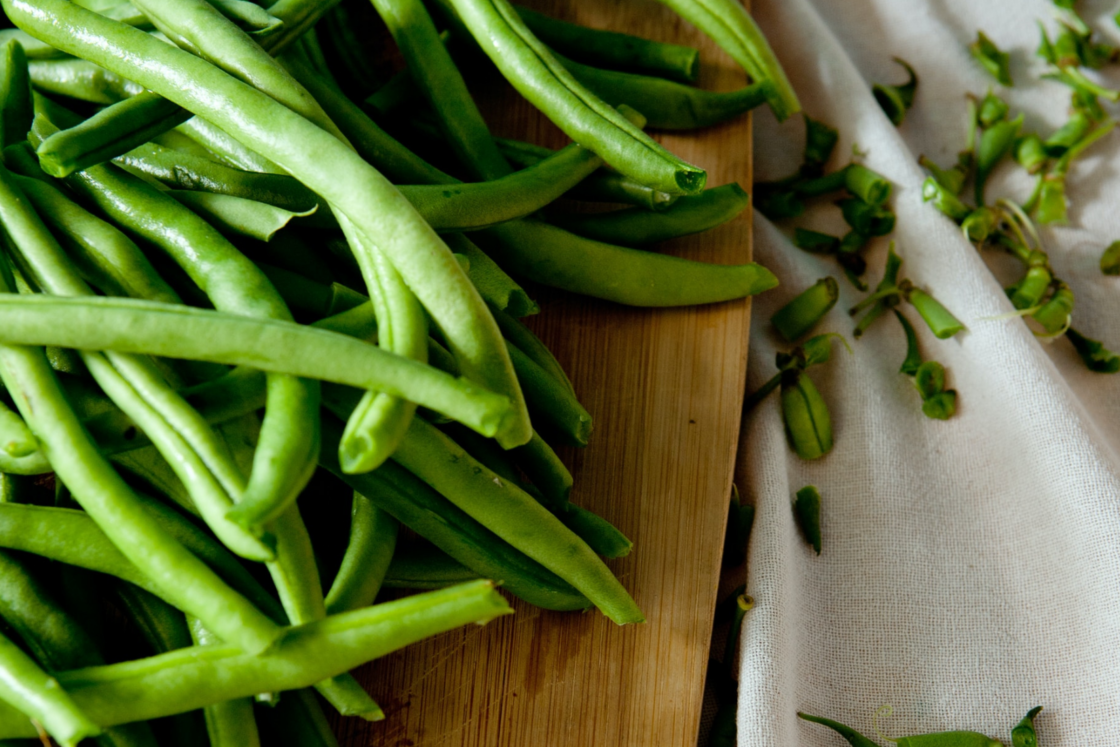Image of a close up of fresh trimmed green beans on a bamboo cutting board with the trimmed ends off to the side on a white table cloth.