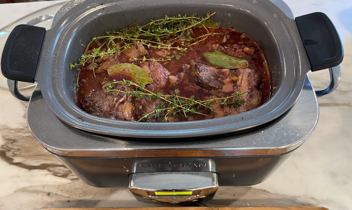 Image of Elite 6-Quart slow cooker used for the best slow cooker review shown filled with braised meat that's been seared over high heat and being cooked in a sauce with fresh thyme and bay leaves on a white marble countertop.