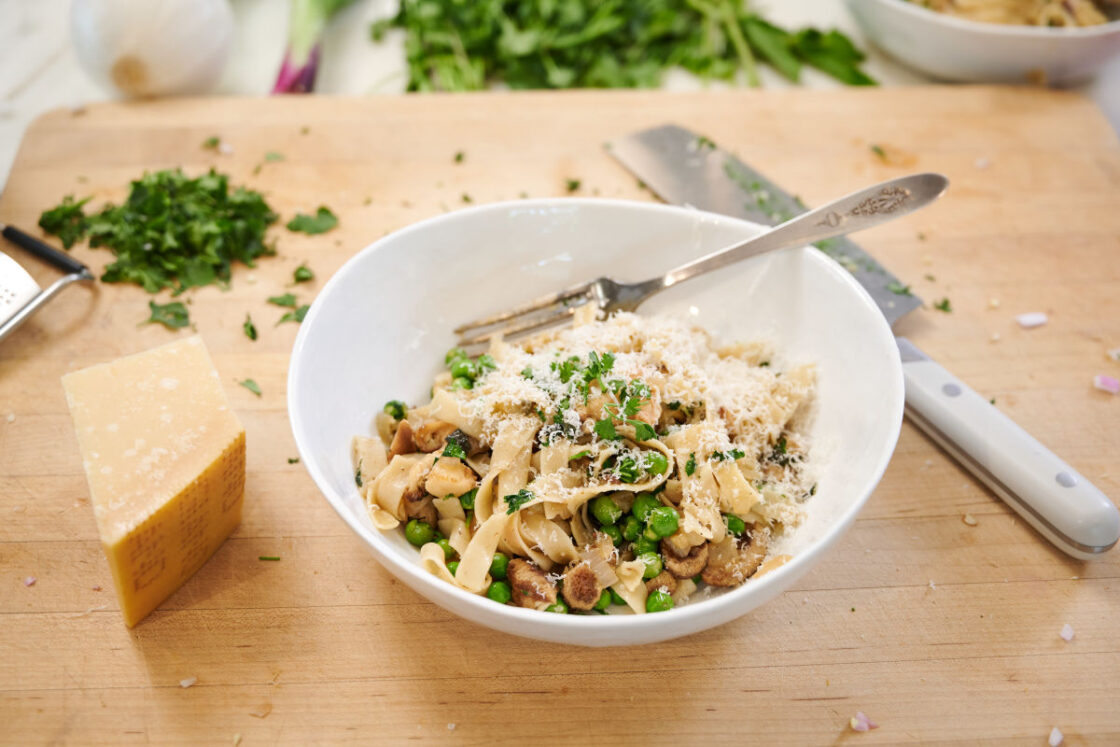 Image of lions mane mushroom recipe with pasta, parmesan cheese, fresh peas, herbs and parmesan cheese in a white pasta bowl on a cutting board with a knife and a block of parmesan cheese and chopped fresh herbs.