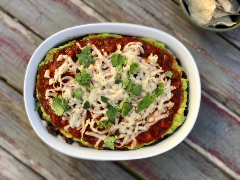 Image of the Healthiest Vegetarian, Vegan 5 Layer Black Bean Dip In All The Land --- final product in a white ceramic baking dish on a wood table via Organic Authority