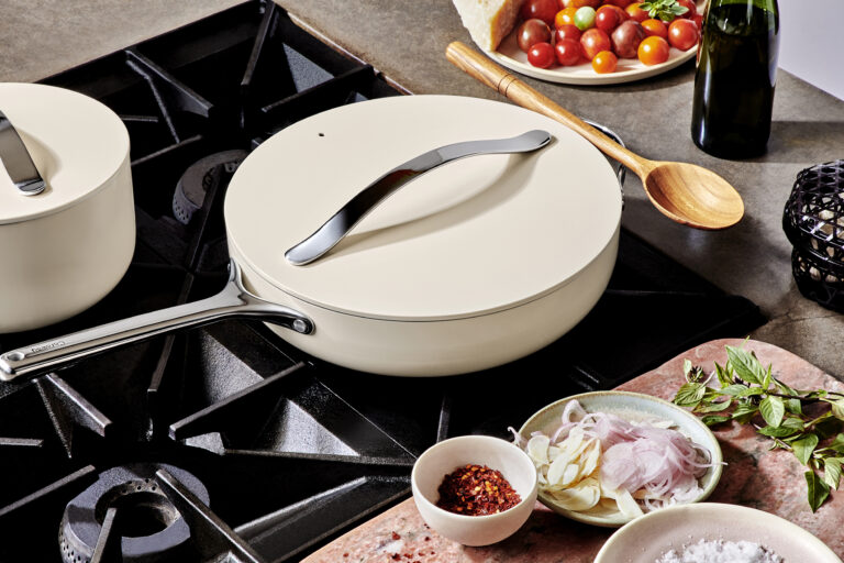 Image of the best ceramic cookware set by Caraway Home on a gas range, a saute pan with lid in cream and a small sauce pot with lid also in cream from the deluxe set. In front of the range is a cutting board with sliced garlic and shallots, a small bowl of crushed red pepper and fresh thai basil.