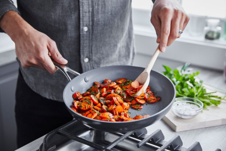 image of GreenPan Valencia Pro one of the best ceramic cookware brands with carrots on range held by man in grey button-down holding wood spoon. In the background there is a wooden cutting board with parsley and a cream sauce in small, clear glass bowl.