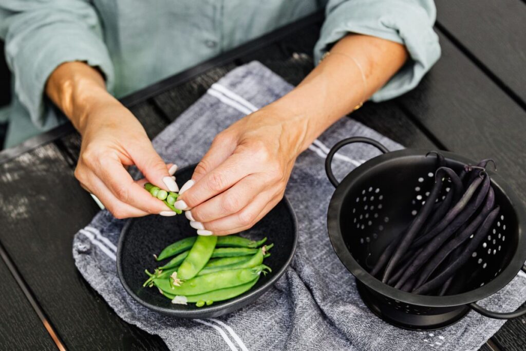 Image of woman's arms/hands with painted white manicured nails shelling fresh peas from their pods over a black bowl on a blue and white towel. Next to the bowl is a black strainer with a dark purple variety of wax beans to prepare with this pasta recipe with lions mane mushroom cooked to golden brown.