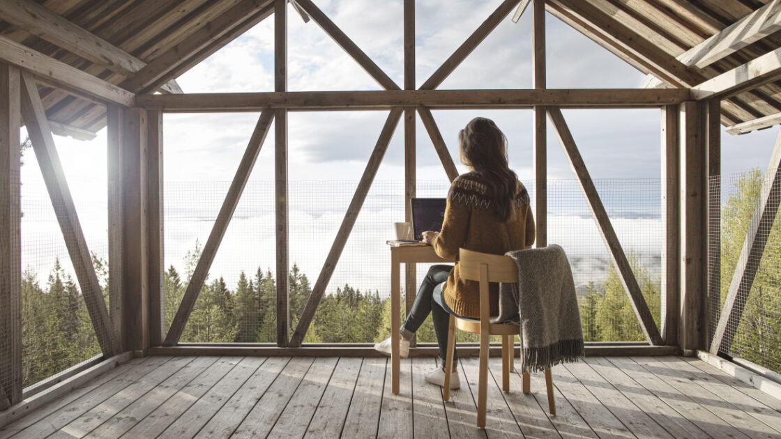 A woman typing on her laptop is sitting at a desk in a room with panoramic view of the treetops.