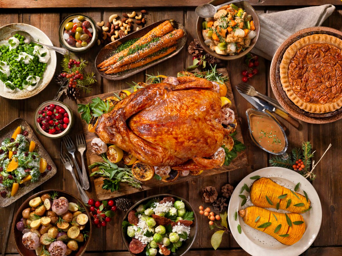 Image of an overhead view of thanksgiving dinner spread on a wood table with turkey, pecan pie, squash, roasted potatoes, salad, and cranberries.