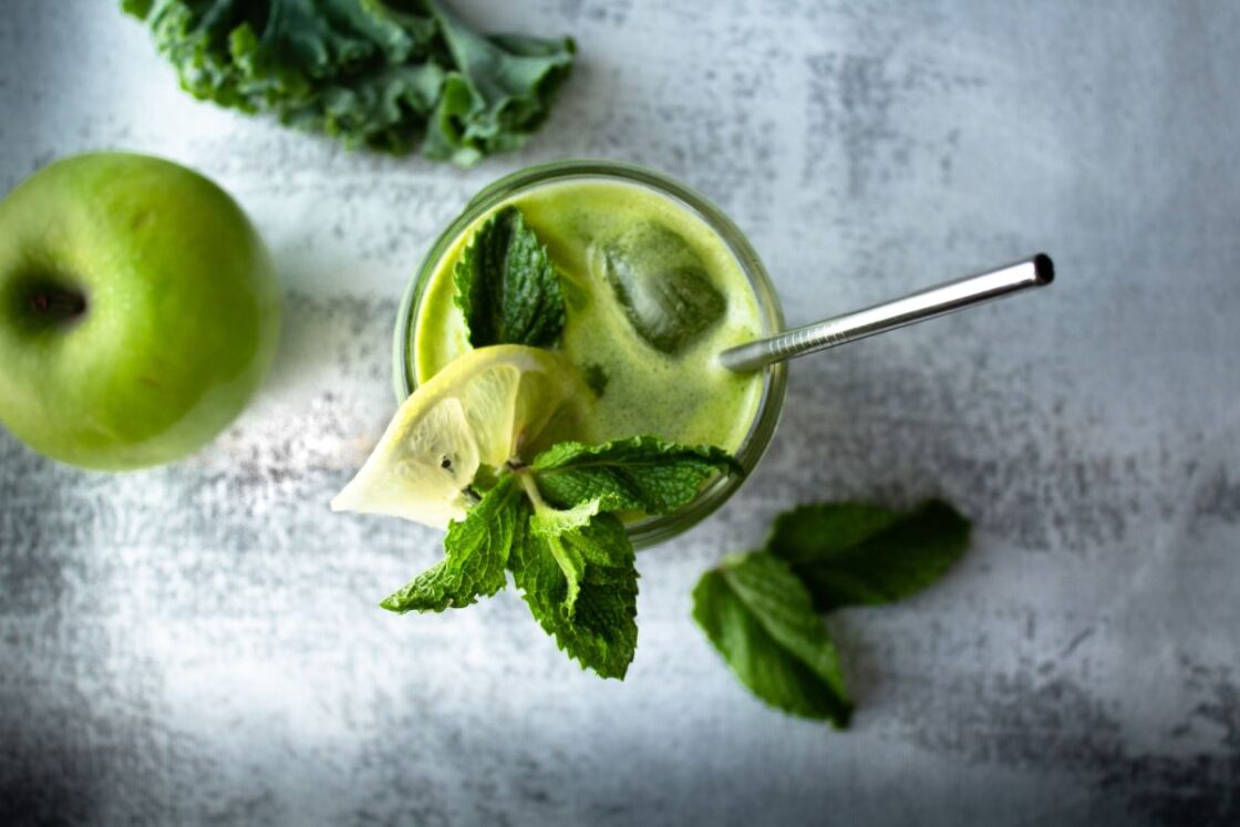 Image of green smoothie or juice combined with wheatgrass powder in glass garnished with mint and lemon and metal straw
