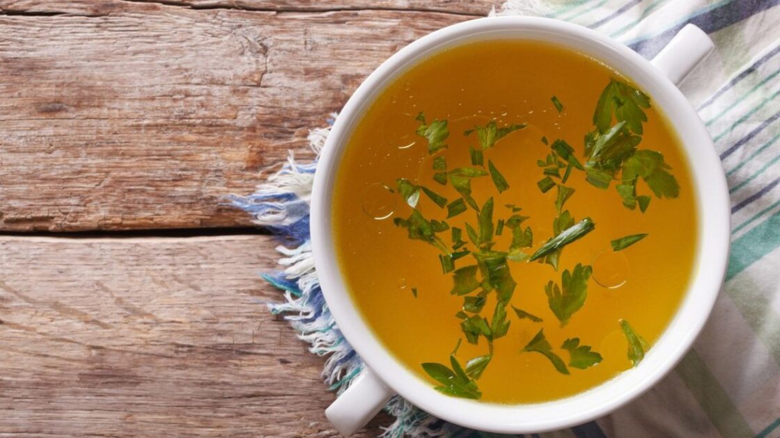 Image of a bone broth recipe in a white bowl with two handles with parsley floating on top on a worn wood table with a striped tea towel.