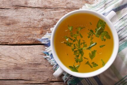 Image of a bone broth recipe in a white bowl with two handles with parsley floating on top on a worn wood table with a striped tea towel.