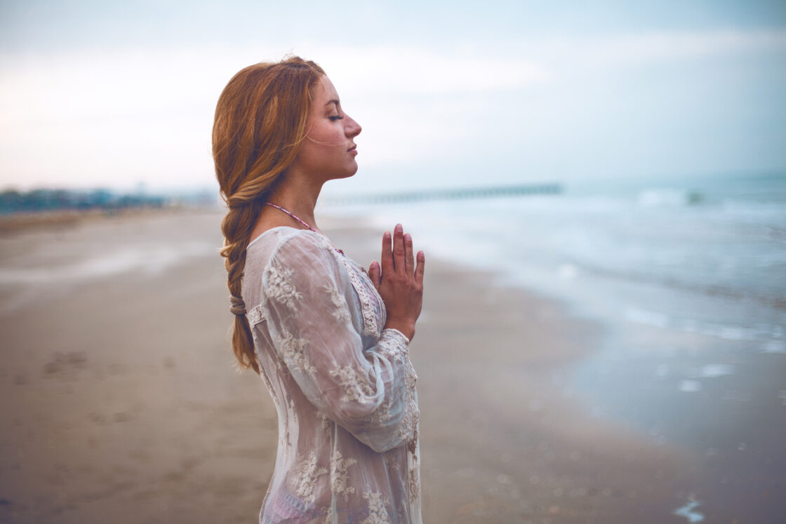 Image of woman standing in prayer pose on the beach, her eyes are closed doing Simple Breathwork Meditation For Beginners