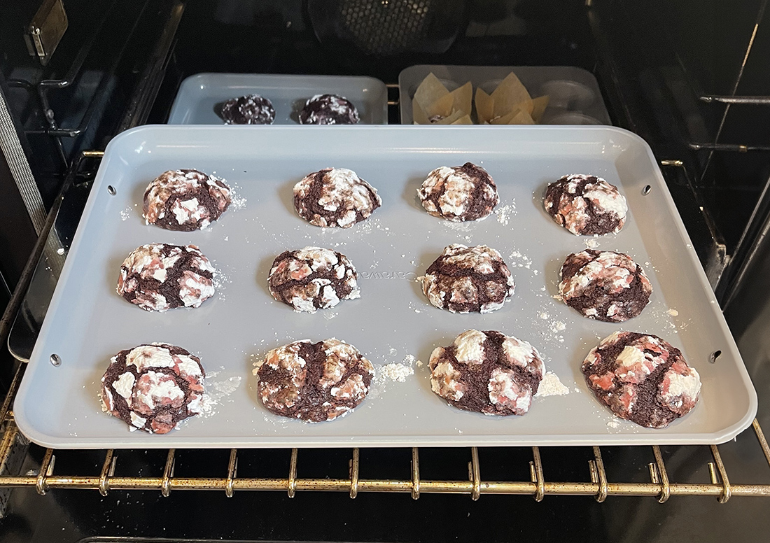 image of inside of oven with Carway baking sheet with one dozen earthquake cookies (chocolate with a crackled powdered sugar crust) pulled out with the top oven rack, a peek of more muffins and cookies being baked beneath via Organic Authority