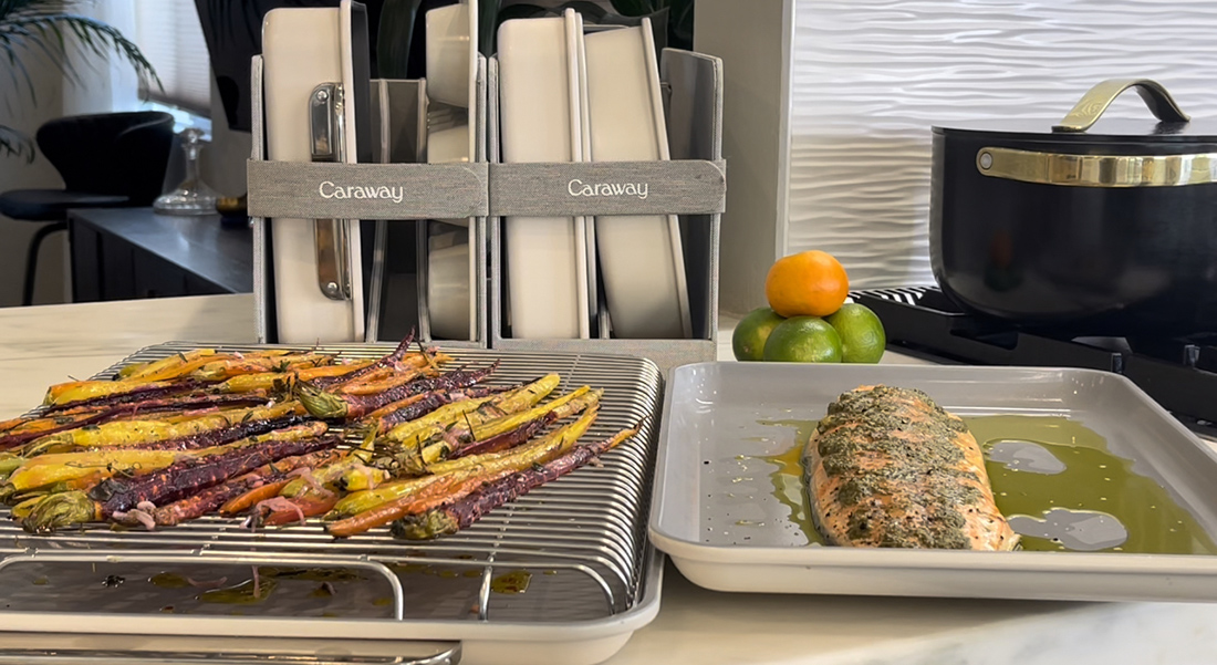 image of a Caraway bakeware sheet pan with a cooling rack on top with colorful roasted carrots drizzled with olive oil, next to that is a pan with roasted salmon with more high-quality olive oil on countertop next to stove with a black covered saucepan. Behind that on the counter rests the Caraway bakeware in white stored storage organizers.