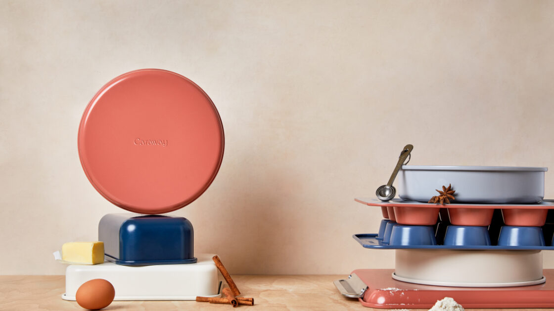 image of Caraway Bakeware Set in mixed colors (perracotta, navy, and white) interestingly stacked in two stacks with a single brown egg, half stick of butter, and sprinkle of flour on the wooden work surface.