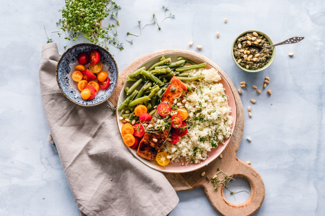 image of a plate of "healthy" food, riced cauliflower, salmon, sliced cherry tomatoes and green beans on a round wooden cutting board next to a speckled blue bowl of sliced cherry tomatoes and a small white bowl of pesto with a silver spoon. Pine nuts and microgreens are sprinkled over the plate and the tablecloth. Even when eating healthy, if there is shame involved it can be just as toxic to our bodies as dieting culture.