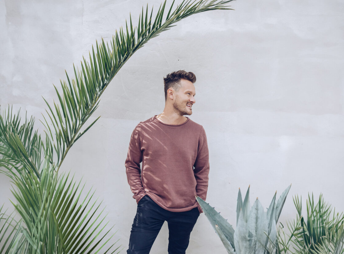 image of Dr. Will Cole standing with head turned to side and hands in pockets wearing worn mauve sweatshirt and dark jeans. Styled image with textured beige background, palm fronds and an aloe plant. Dr. Cole guides away from toxic diet culture.