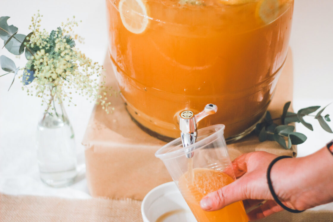 image of large glass vessel with pour spout holding orange liquid with sliced fruit. Hand is holding a plastic cup beneath, catching the flowing orange liquid for full body cleanse.