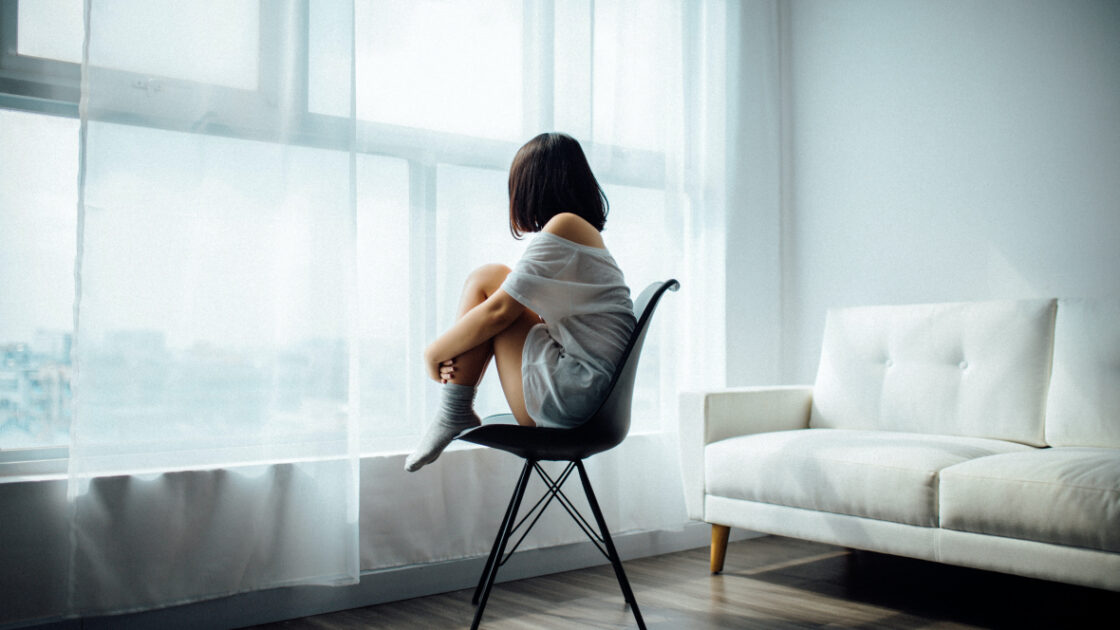 image of pensive woman sitting in chair looking out window through sheer, white curtains, next to white sofa wondering if she needs a full body cleanse.