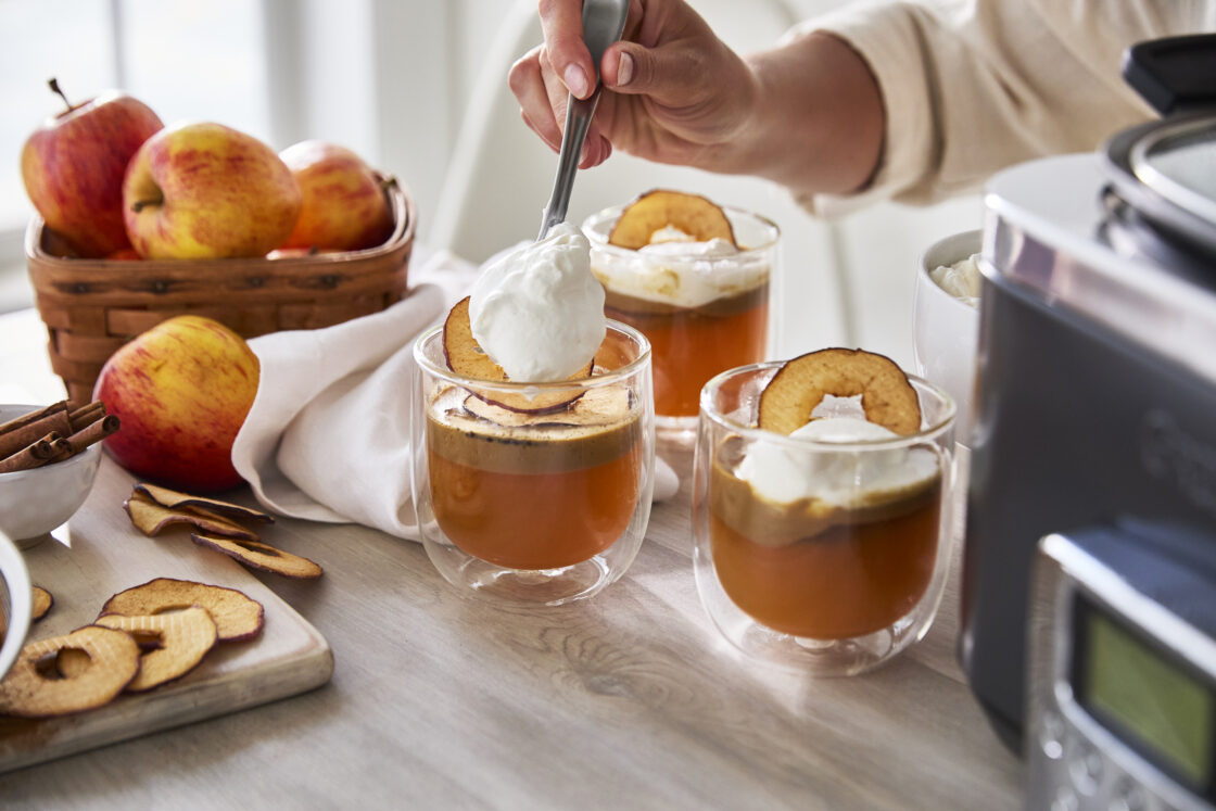 Image of a GreenPan Slow Cooker and three clear glass double walled glasses filled with apple cider and a dollop of whipped cream is being added to one. On a wood surface with a wood cutting board and sliced apples and a basket of apples.