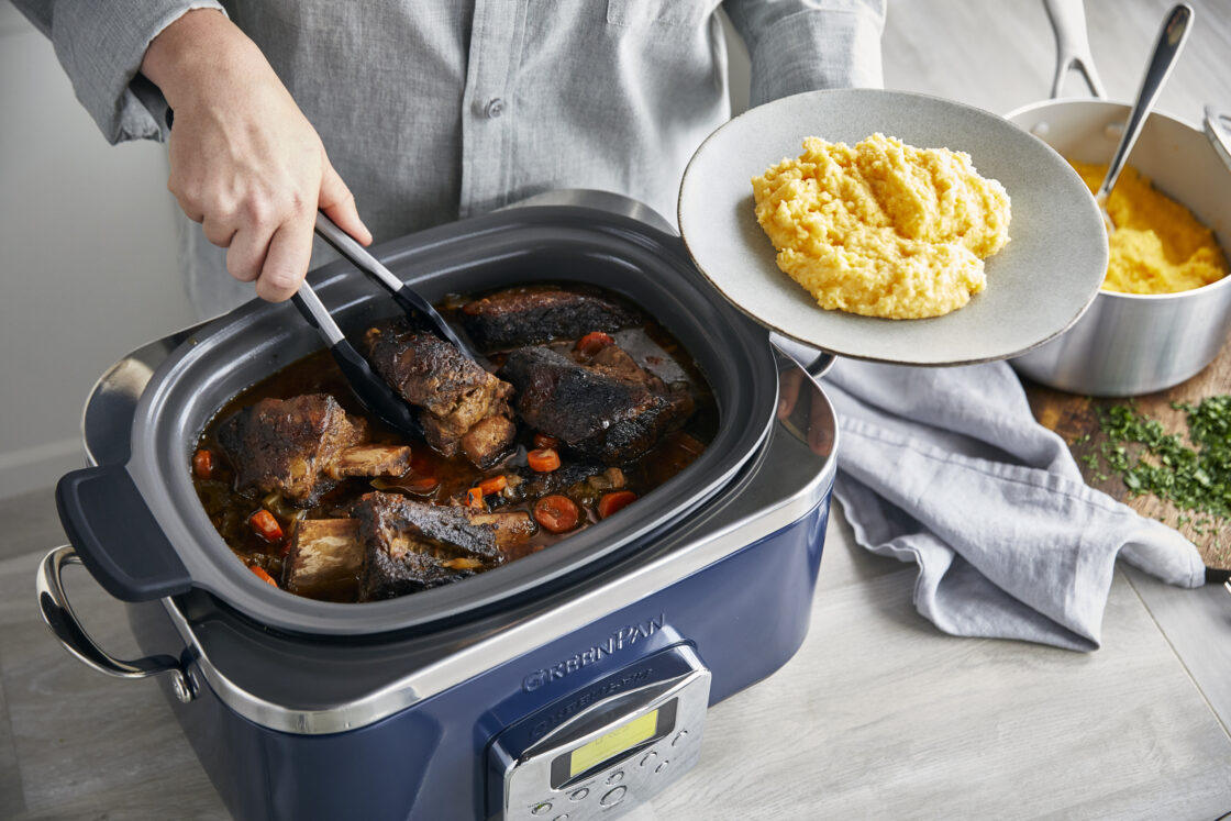 Image of an overhead shot of the best slow cooker, a GreenPan 6-Quart slow cooker in navy, with seared and stewed meat being grabbed by tongs and served over polenta on a wood surface.