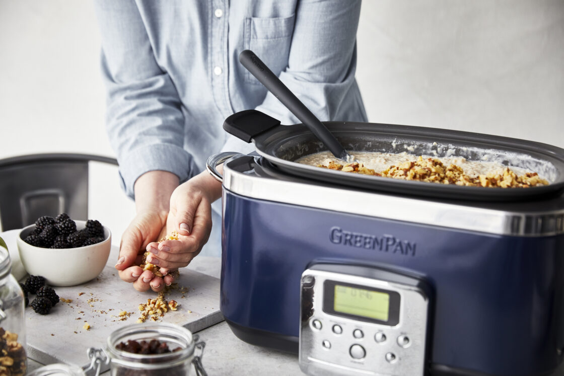 Image of the best 6-quart slow cooker in navy filled with maple brown sugar oatmeal, cook stands behind scooping up walnuts to add to the top, a small white bowl of blackberries sits on the counter.