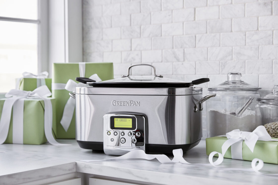 Image of a GreenPan Slow Cooker in a stainless steel finish looking sleek on a white marble countertop with a white marble backsplash. Styled alongside is a curling white ribbon, several wrapped packages in a mint green with white ribbons, and 2 glass canisiters with beans and flour.
