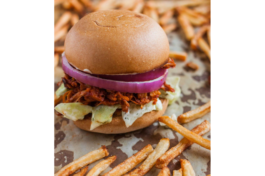 Image of a vegan bbq jackfruit pulled pork slider with slaw on the bottom, bbq jackfruit "pulled pork," red onion, on a bun. All on a sheet pan with parchment paper, with french fries scattered around.