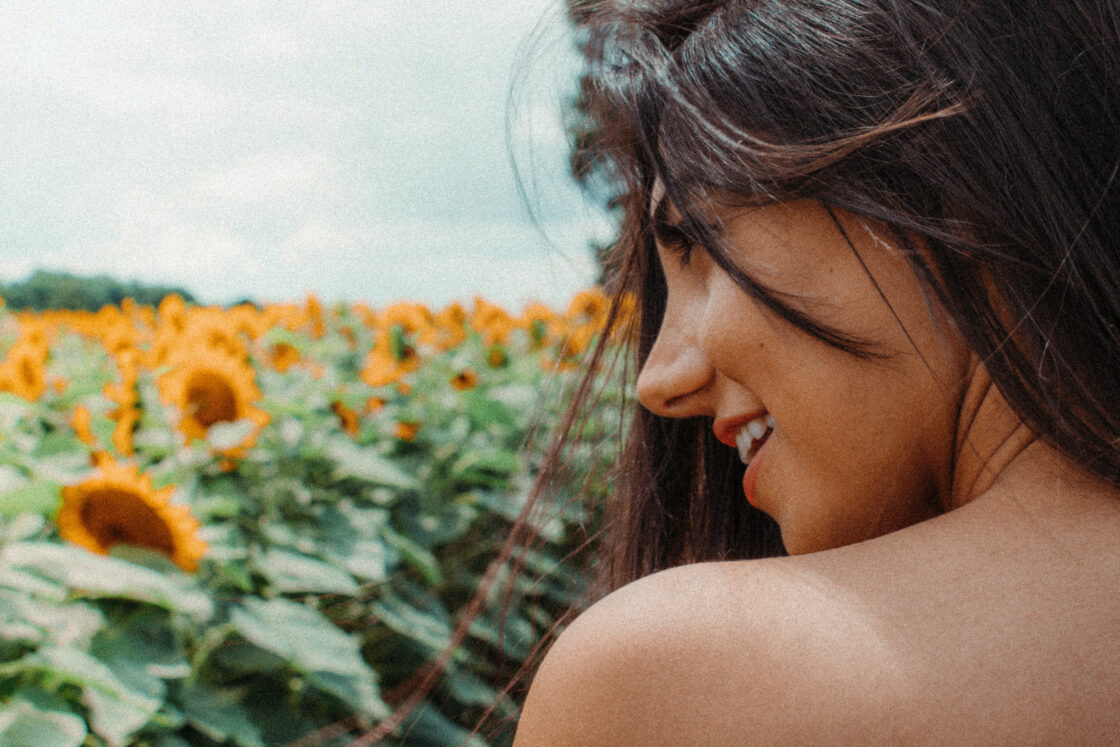 Image of woman smiling in profile looking over bare shoulder with field of sunflowers in the background showing healthy skin via Organic Authority. Liquid chlorophyll benefits may help improve skin (science is thin however).