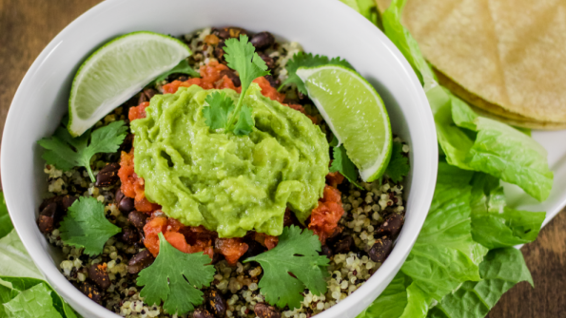 Image of Burrito Bowl with Spiced Black Beans and Quinoa Recipe served in a white bowl on a white platter with lettuce and corn tortillas on a wood table.