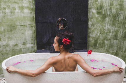 Image of a woman in a modern, stone bath, sitting up with arms splayed to each side facing a wall with bath fixture. She wears a hibiscus flower in her hair and shows the benefits of Epsom salt baths. Why to Take an Epsom Salt Bath