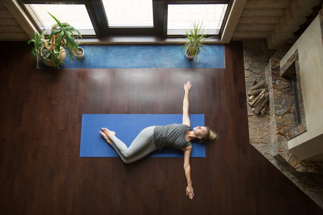 Attractive young woman working out in living room, doing yoga exercise on wooden floor, lying in Belly Twist Pose, Jathara Parivartanasana, resting after practice, full length, top view