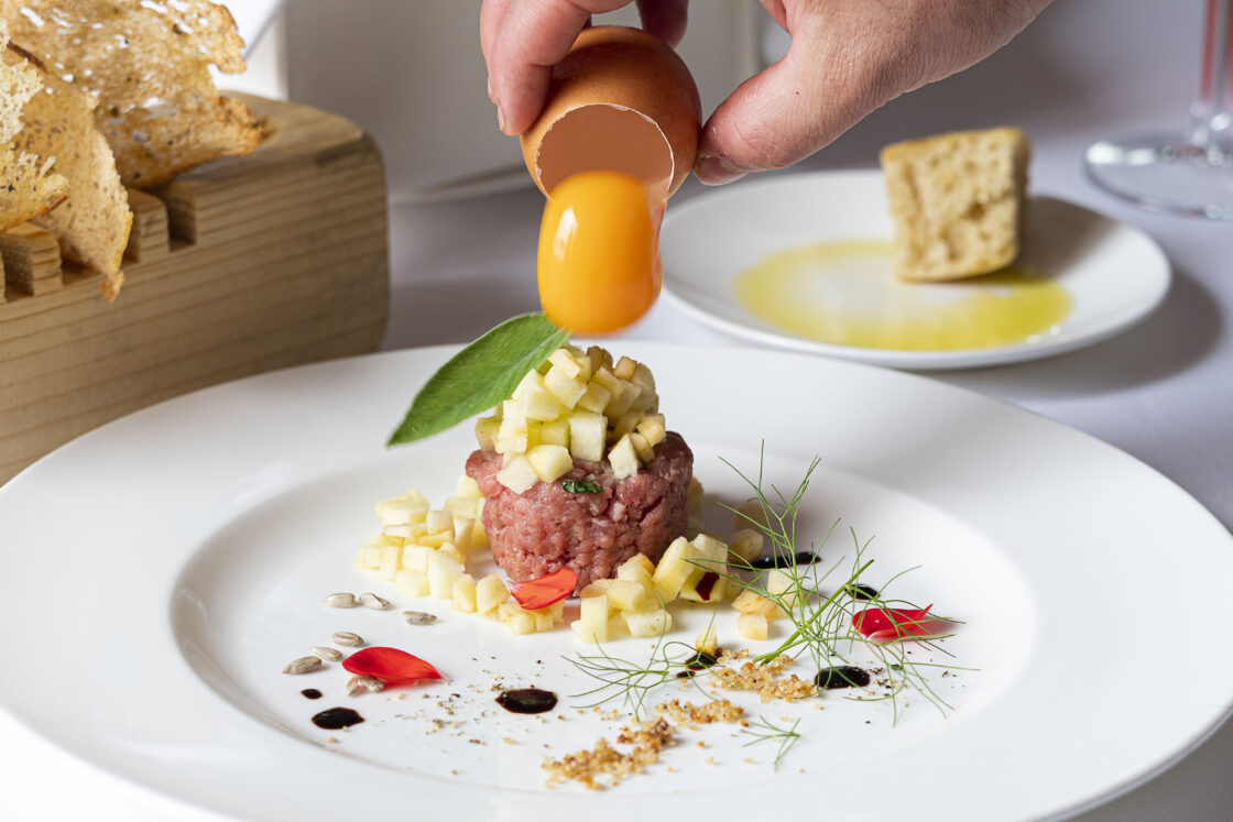 Image of a plate of steak tartare with a quail egg yolk being placed onto the top straight out of the shell. This is one of the many life-changing experiences you can enjoy at this Mount Etna wellness vacation.