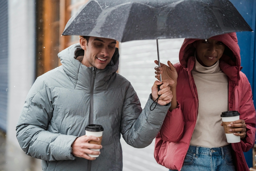 Image of a smiling young couple walking in the rain with several consumer products that could contain PFAS: raincoats, umbrellas, and takeaway coffee cups.