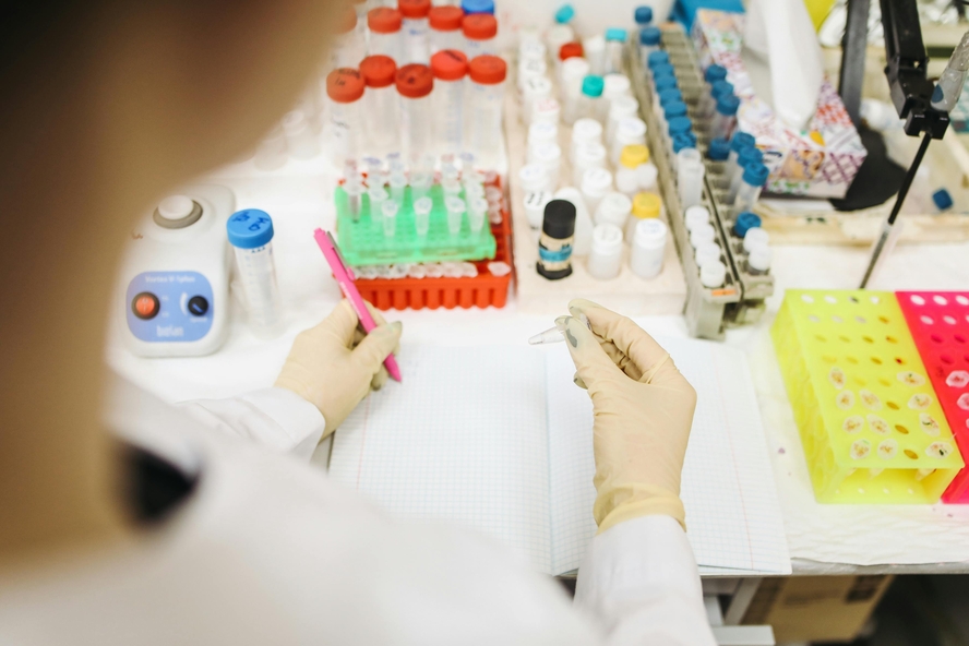 Image of a scientist in a lab coat and gloves holding a test tube, surrounded by chemical equipment, and writing in a notebook.