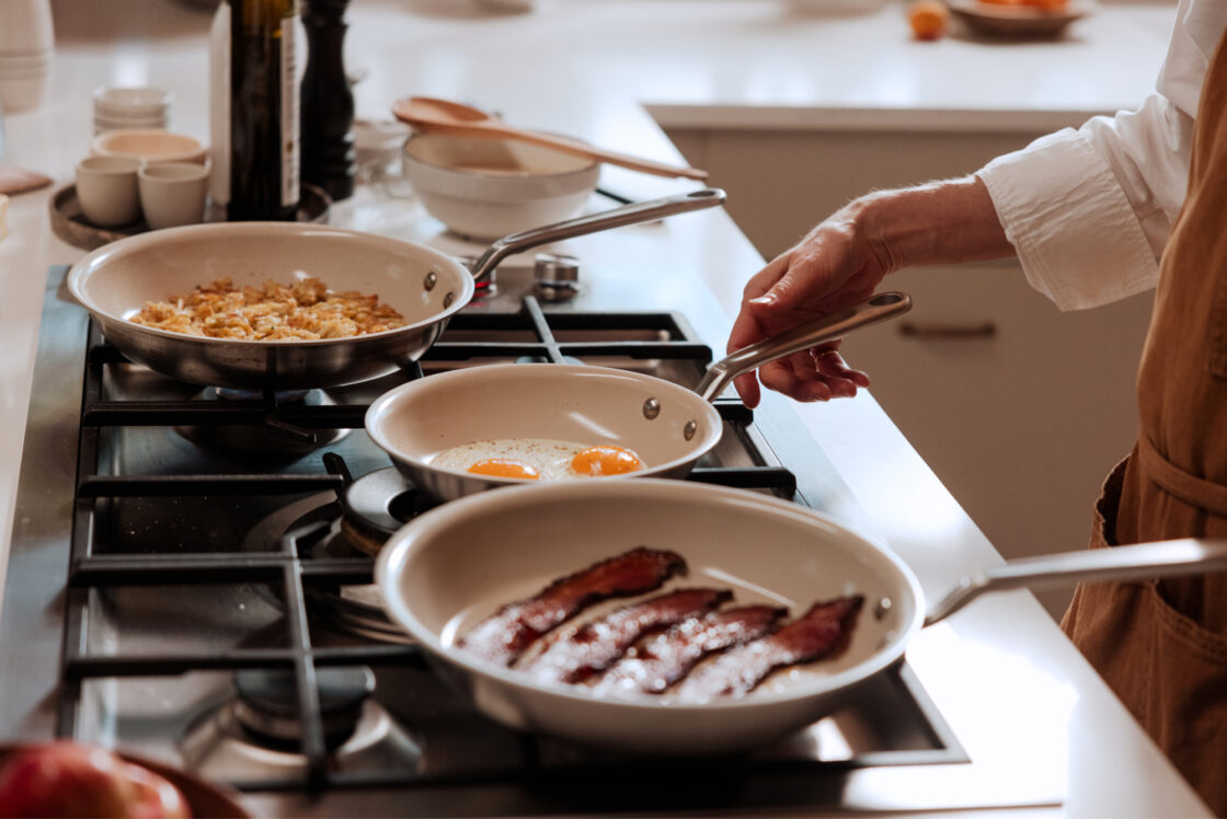 Image of Made In nonstick cookware on a stove top with one pan filled with sizzling bacon, one with fried eggs, and the other with potatoes. 