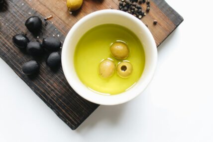 Image of an overhead view of a ramekin of oiive oil with 3 green olives on a cutting board with other olives, raising the question, Is high phenolic olive oil better for you than good old EVOO?