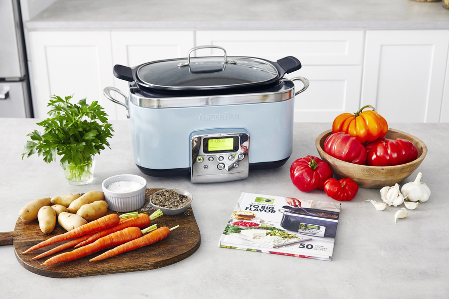 Image of a GreenPan Elite 6-Qt. Slow Cooker in Blue Haze, on a countertop with a cookbook, a bowl of heirloom tomatoes, a cutting board with potatoes and carrots, and other cooking ingredients.