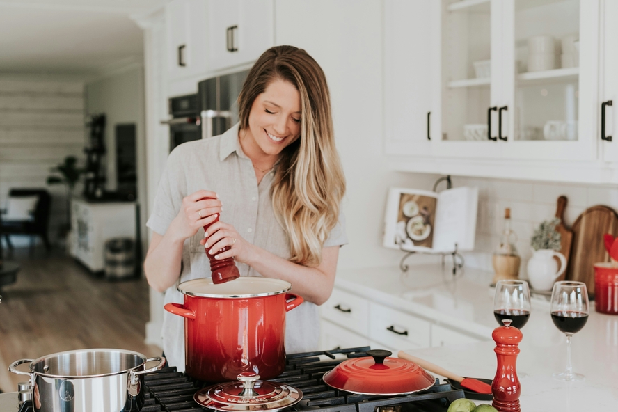 Image of woman grinding pepper over a red ceramic pot on the stove in a beautiful kitchen.