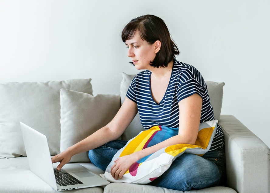 An image of a woman sitting on a grey couch with a pillow on her lap, looking at a laptop set beside her while having a virtual meeting with her nutritionist.