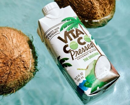 Image of a carton of Vita Coco Pressed Coconut Water floating in a swimming pool with two brown coconuts.