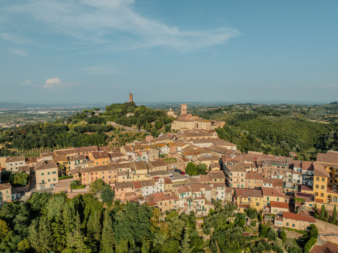 Image of the village of San Miniato shown from above, with terracotta colored buildings surrounded by greenery.