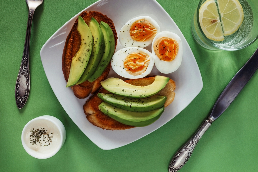 Image of a plate with toast topped with avocado and eggs, on a green background alongside a glass of lemon water.