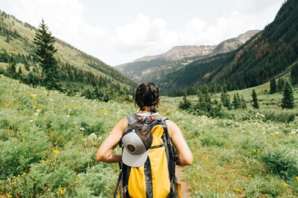 Image of a woman on a hike, wearing a yellow backpack and standing in front of an expansive green field with a view of mountains in the distance.