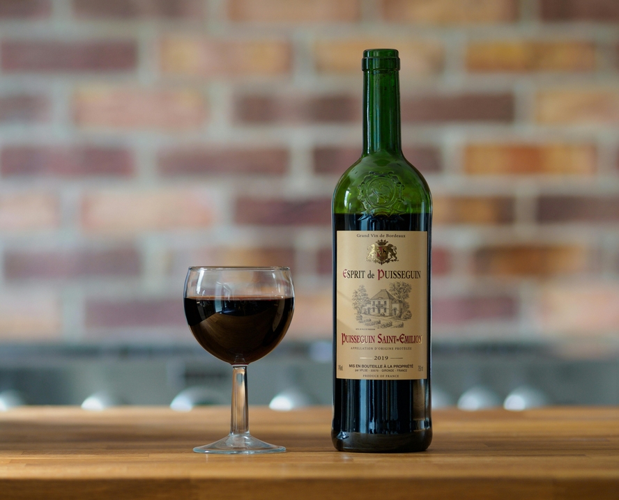 Image of a bottle of red wine with a brown label, sitting next to a full glass of the same wine on the table.