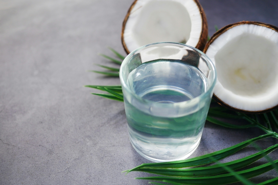 Image of a clear glass of coconut water, with two halves of a brown coconut behind it.