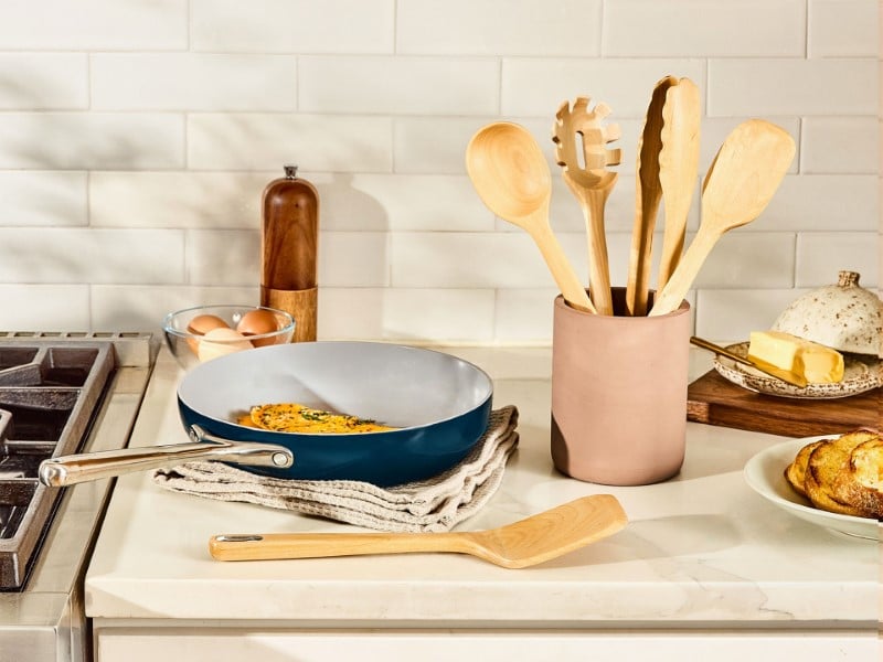 Image of a bright kitchen with Caraway cookware and Caraway birch wood utensils spread out with an array of food.