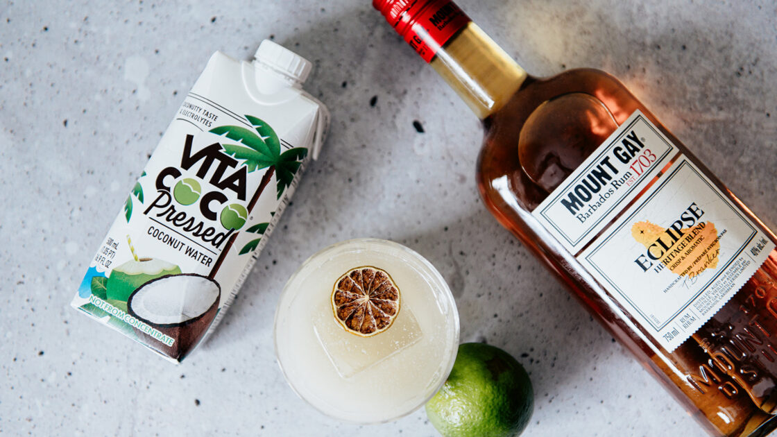 Image of a flat-lay of cocktail ingredients for coconut water drinks, from a carton of Vita Coco Pressed Coconut Water to a bottle of Mount Gay Rum, set next to a refreshing cocktail.