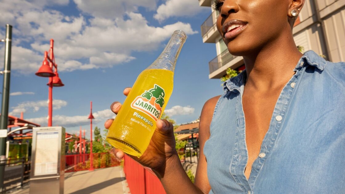 Image of a woman standing on a train platform in a denim vest top, holding a yellow sugar-sweetened soda beverage.