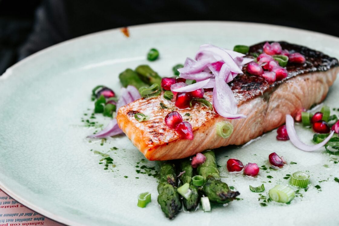 Image of a beautiful salmon filet, which would fit right into the best diet for brain health. on a white plate and arranged with asparagus, onions, and pomegranate seeds.